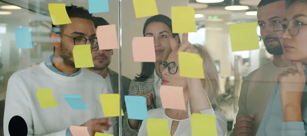 A group of people gather around a clear wall with post it notes.     