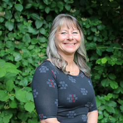 A woman smiling with a background of green bushes.