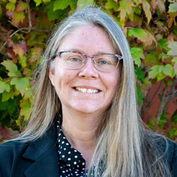 A woman with long gray hair and glasses smiles.